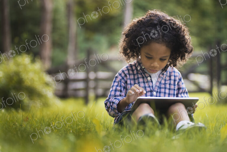 A small boy sitting on the grass using a tablet computer.