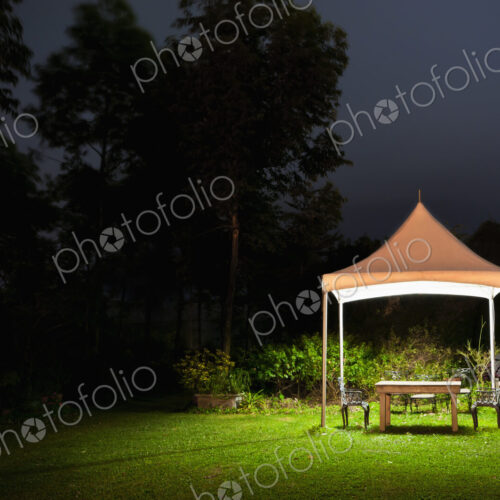 A small gazebo in a garden, a table and seat and lights at night