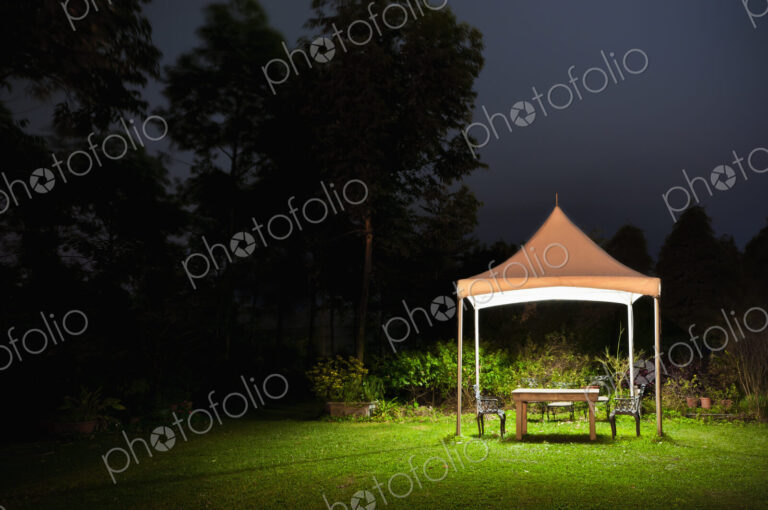 A small gazebo in a garden, a table and seat and lights at night