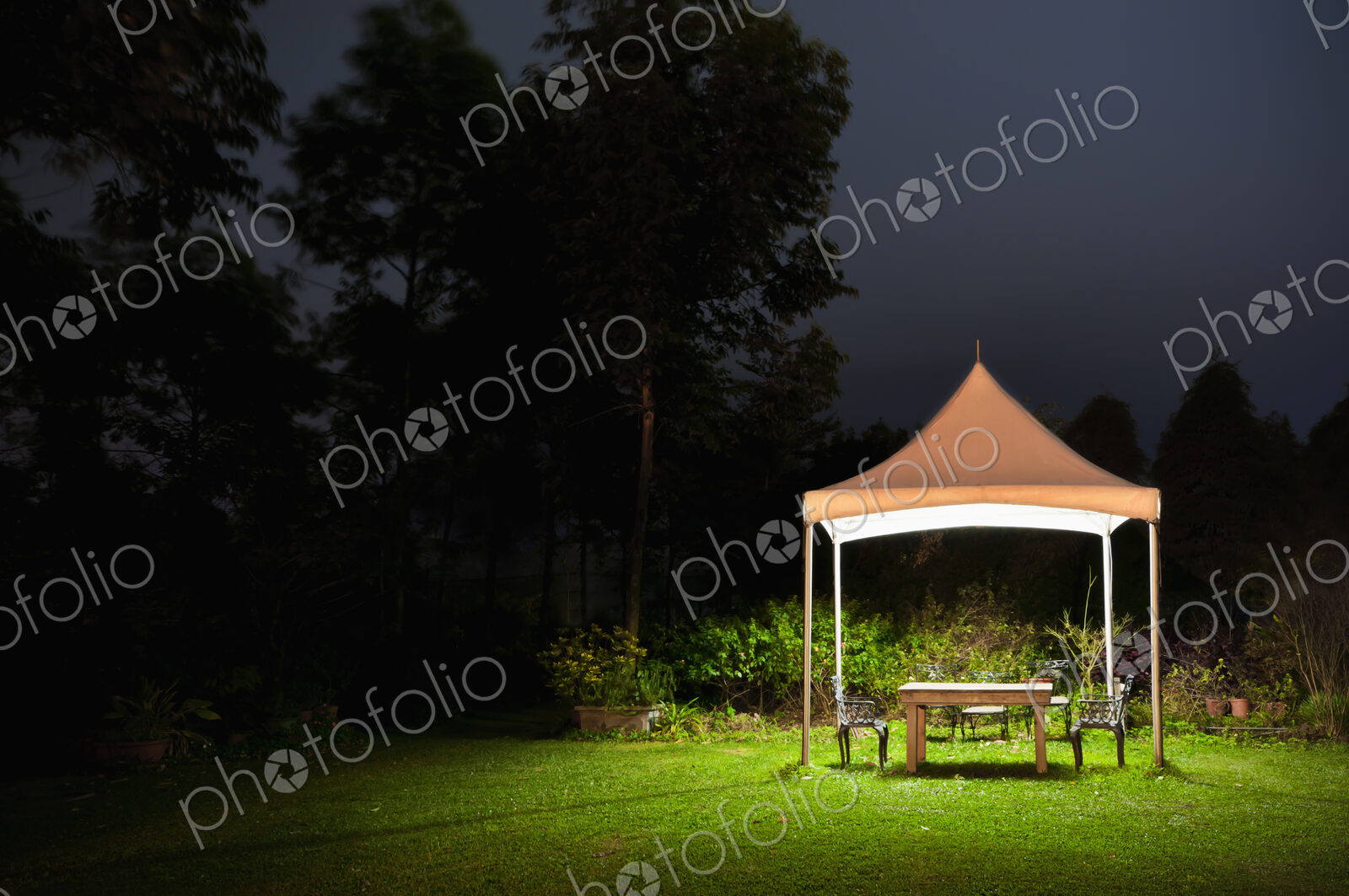 A small gazebo in a garden, a table and seat and lights at night