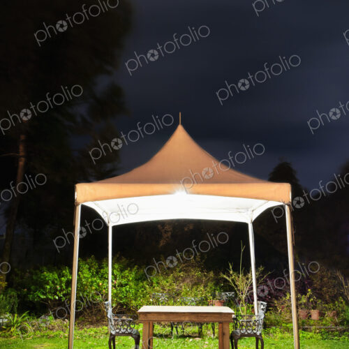 A small gazebo in a garden, a table and seat and lights at night