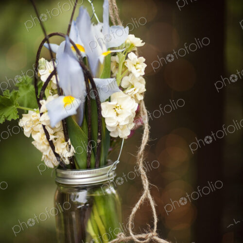 A small glass jar hanging from a wire, with iris and scented stock flowers. A floral…