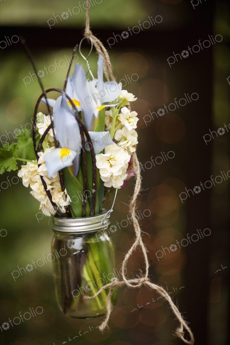A small glass jar hanging from a wire, with iris and scented stock flowers. A floral…