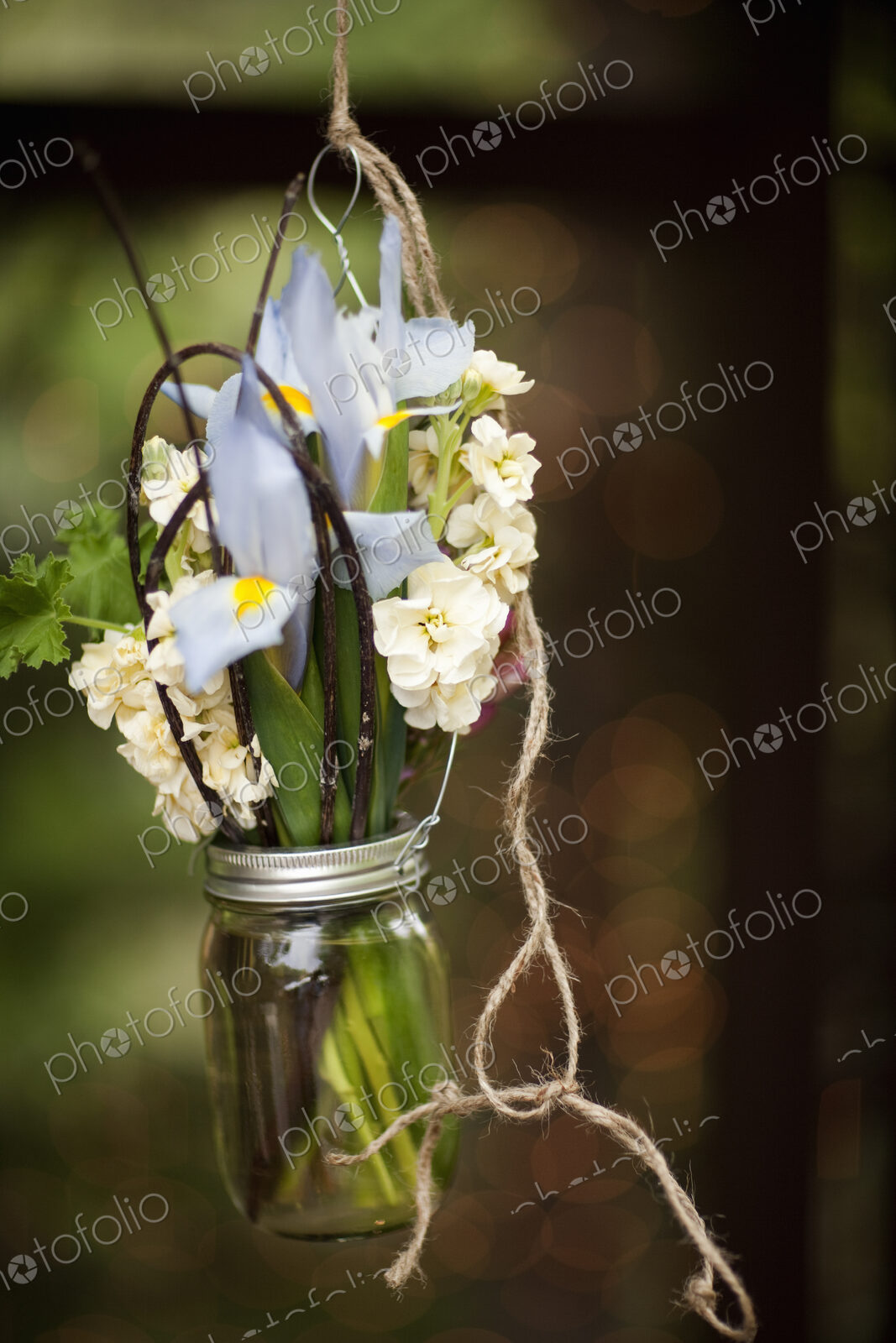 A small glass jar hanging from a wire, with iris and scented stock flowers. A floral…