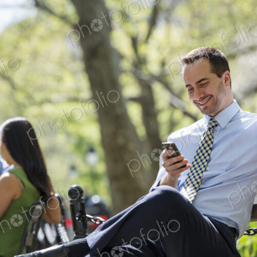 A small group of people. A couple on a bench in the background, and a man checking his… A small group of people. A couple on a bench in the background, and a man checking his…