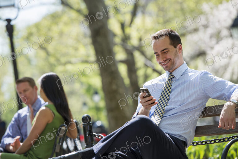 A small group of people. A couple on a bench in the background, and a man checking his…