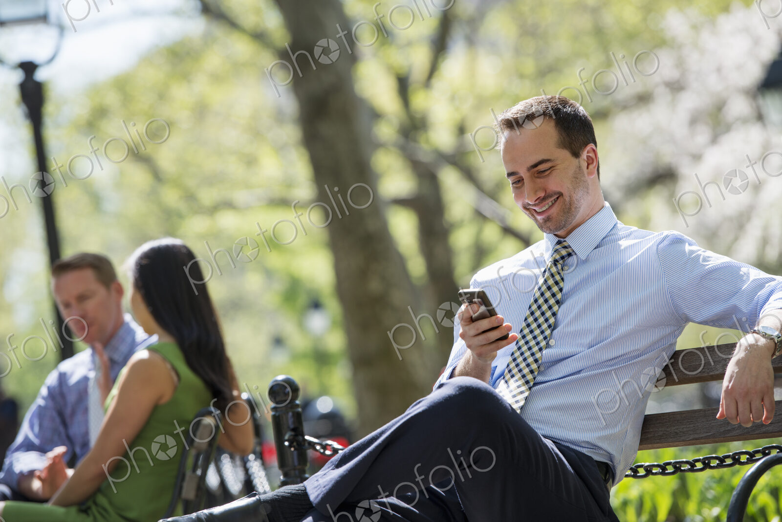 A small group of people. A couple on a bench in the background, and a man checking his…