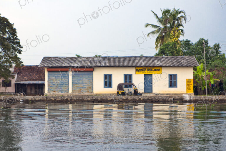 A small house by a waterway with a tuk tuk taxi in front, in the backwaters of Kerala,…