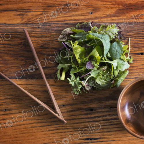 A small round polished wooden bowl and a clutch of organic mixed salad leaves, with…