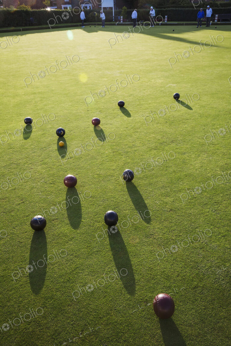 A small yellow target jack ball and a cluster of lawn bowls around it. People in the…