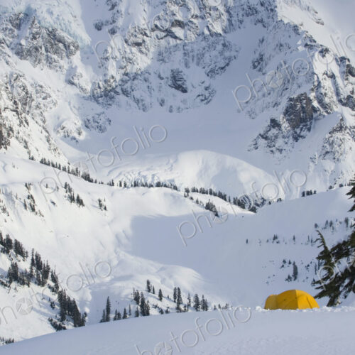A small yellow tent pitched in deep snow on a slope, view of the steep slopes of the… A small yellow tent pitched in deep snow on a slope, view of the steep slopes of the…