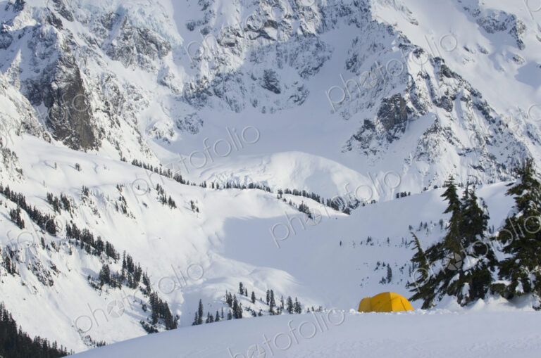A small yellow tent pitched in deep snow on a slope, view of the steep slopes of the…