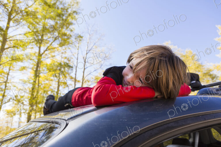 A smiling 6 year old boy lying on the hood of a blue SUV in woodland.