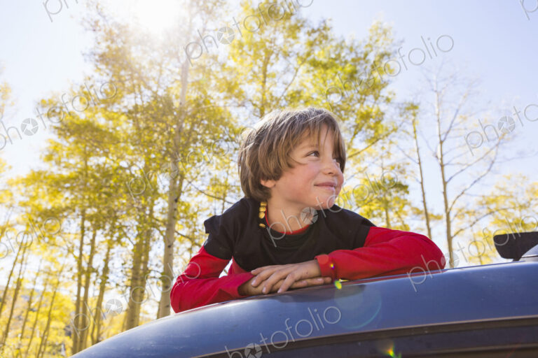 A smiling 6 year old boy lying on the hood of a blue SUV in woodland.
