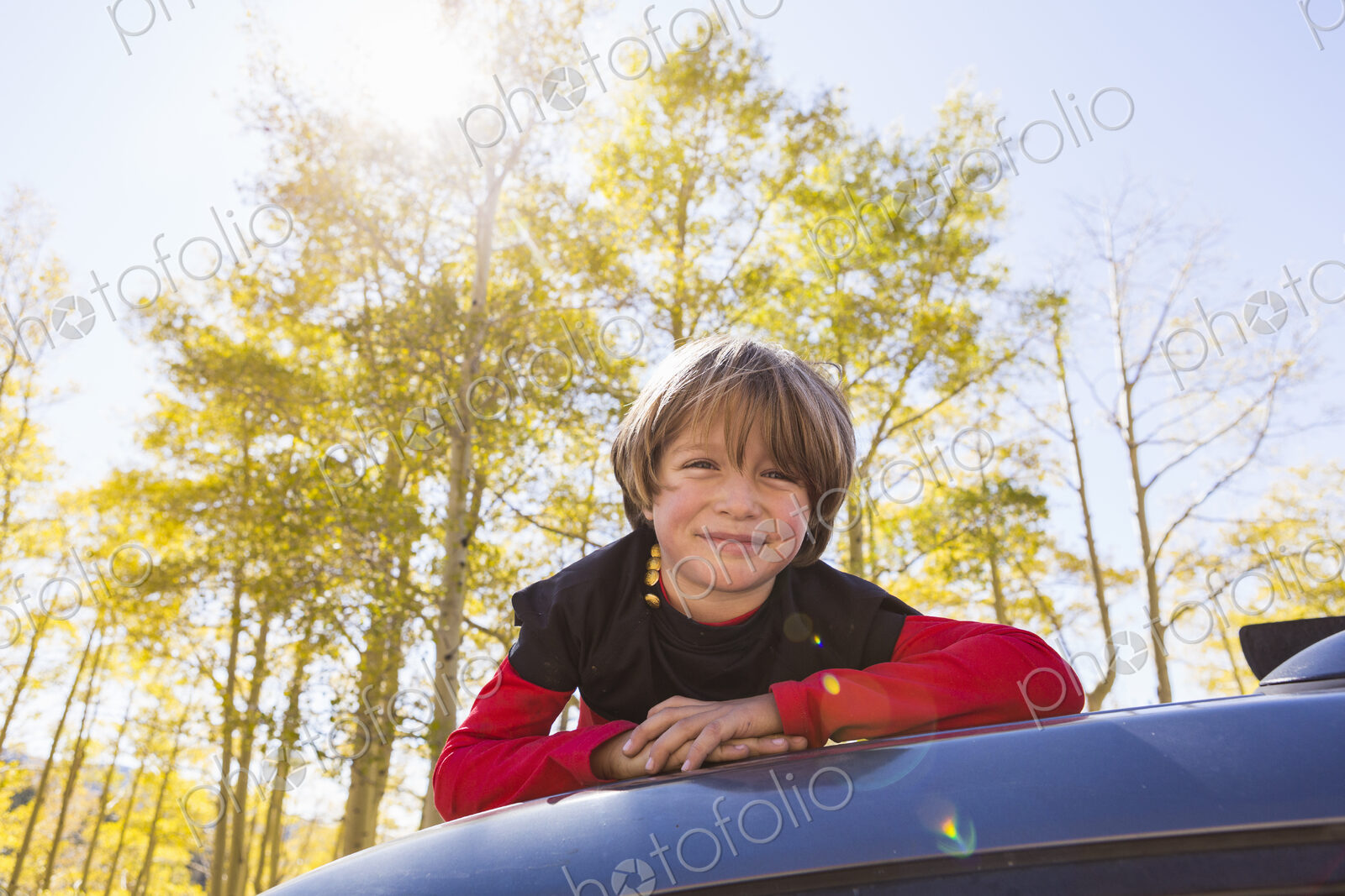 A smiling 6 year old boy lying on the hood of a blue SUV in woodland.