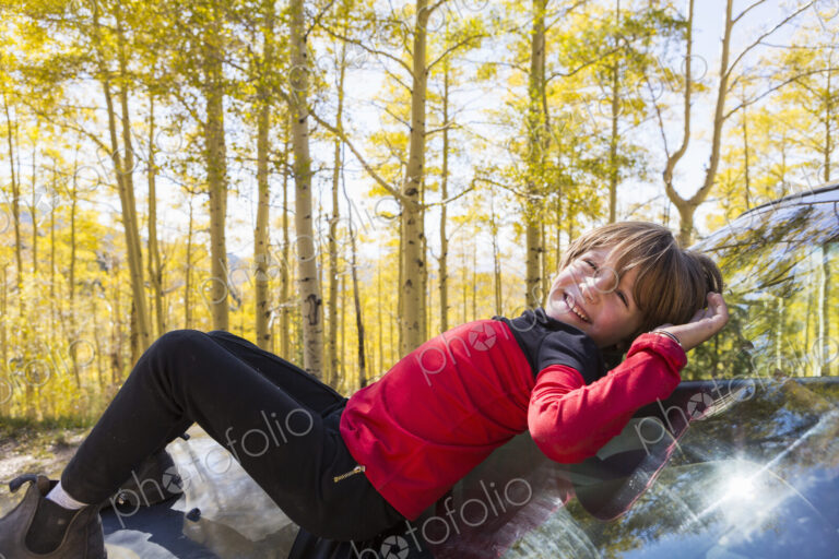 A smiling 6 year old boy lying on the hood of a blue SUV in woodland.
