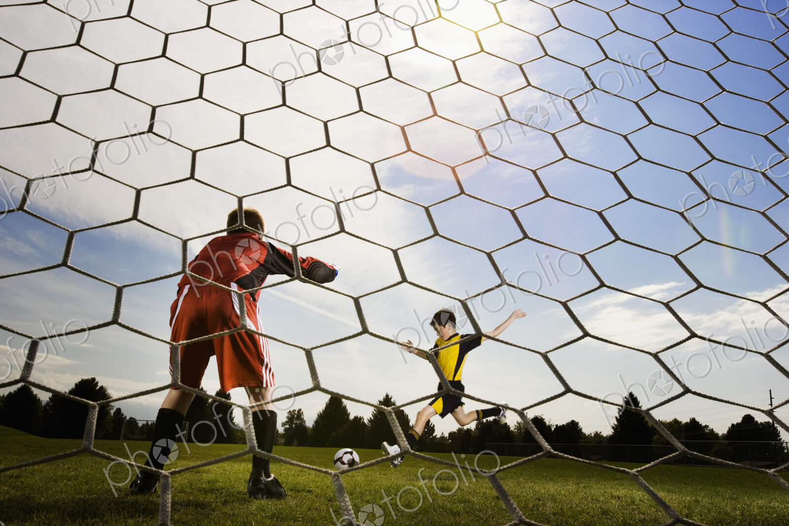A soccer player taking aim and kicking at the goal, view from behind the goal keeper.