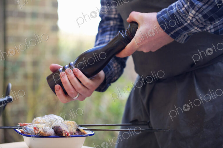 A spatchcocked game bird being seasoned for cooking, a man's hand grinding pepper in a…