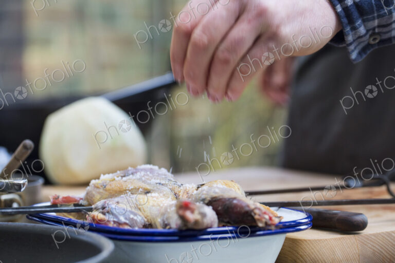 A spatchcocked game bird being seasoned for cooking, a man's hand sprinkling salt on…