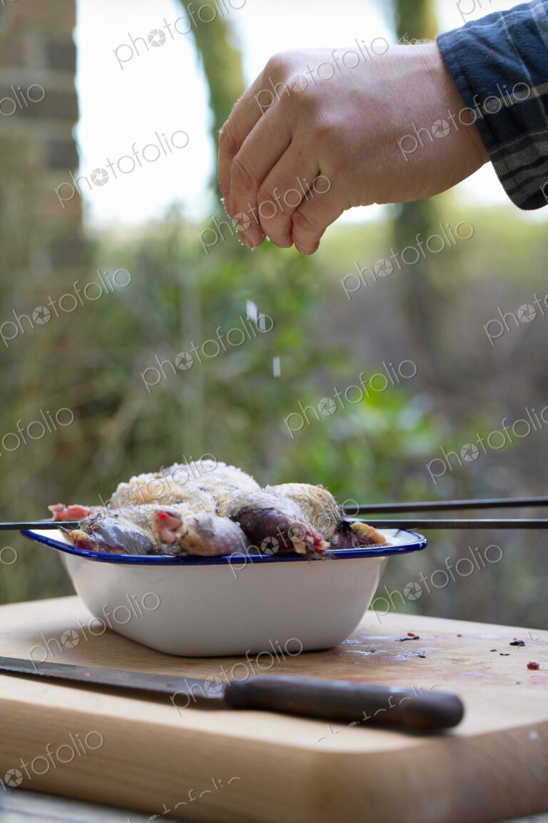 A spatchcocked game bird being seasoned for cooking, a man's hand sprinkling salt on…