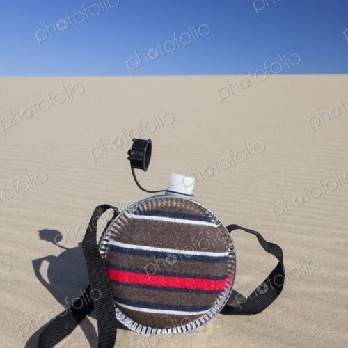 A striped fabric-covered canteen with a shoulder strap rests on the rippled surface of… A striped fabric-covered canteen with a shoulder strap rests on the rippled surface of…
