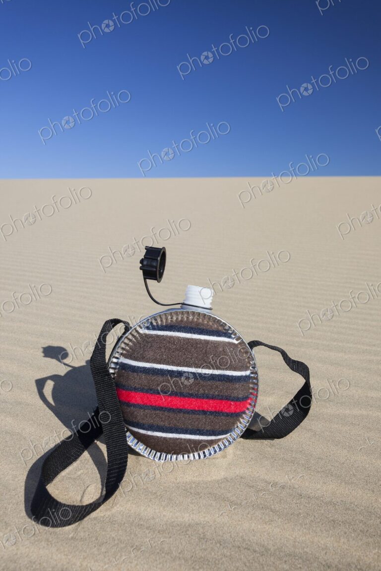 A striped fabric-covered canteen with a shoulder strap rests on the rippled surface of…