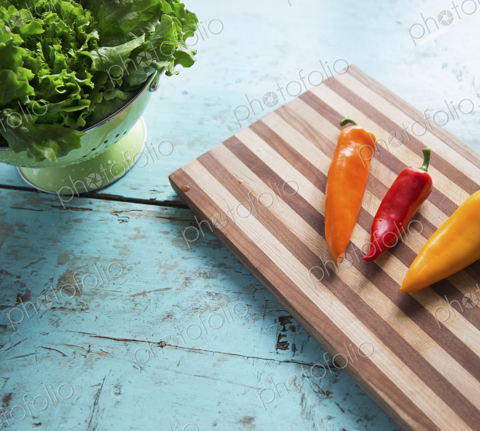 A tabletop with a wooden chopping board and three sweet peppers, and a bowl of salad…