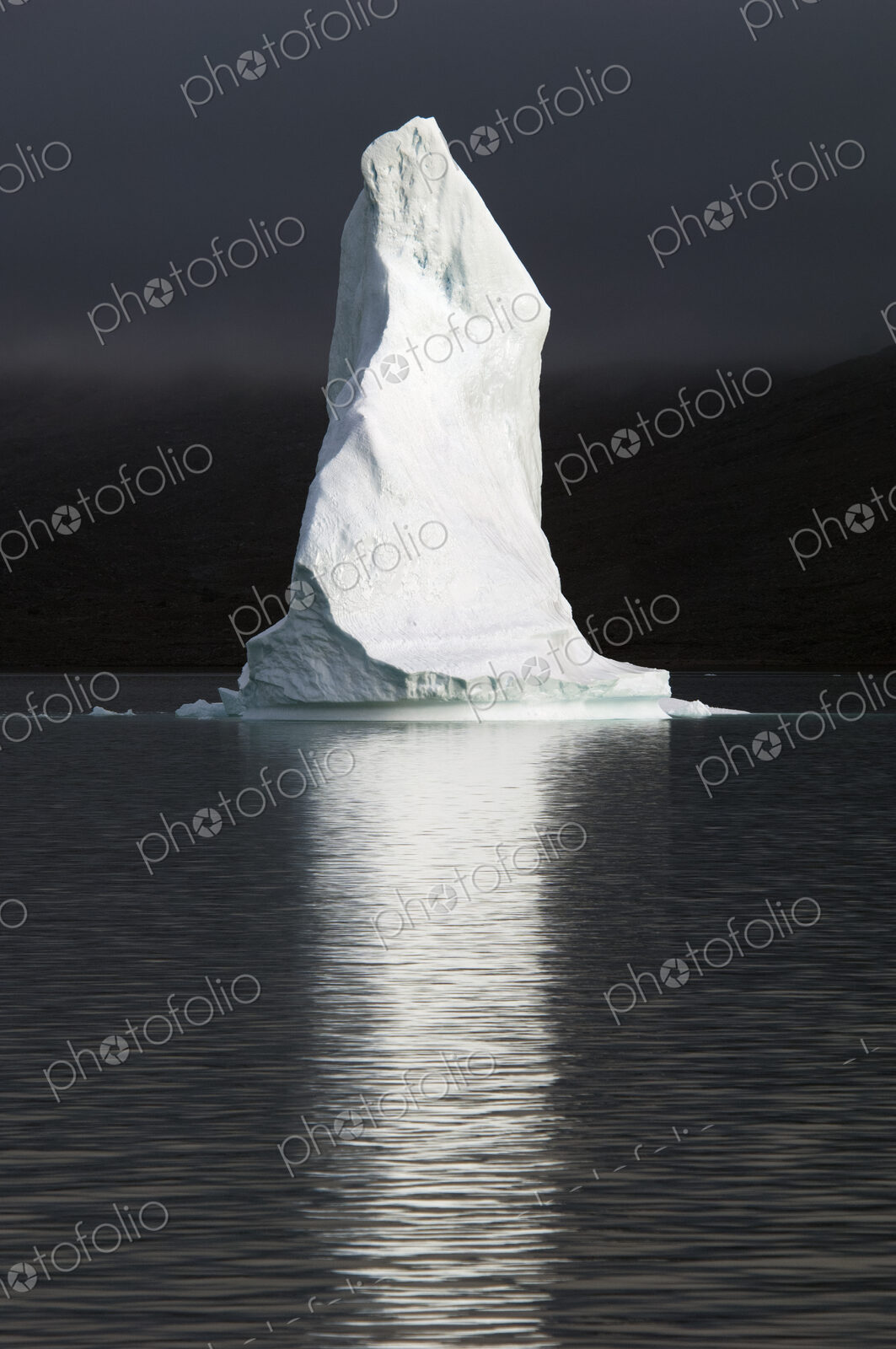 A tall pillar of ice, the top of an iceberg lit up floating on the sea, reflected in…