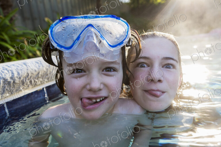 A teenage girl and brother in a warm pool, looking at the camera, sticking their…
