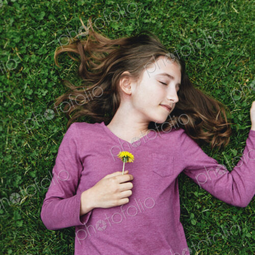 A ten year old girl lying on the grass with her eyes closed, holding a dandelion flower.