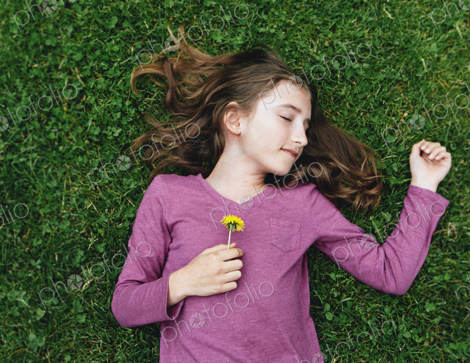 A ten year old girl lying on the grass with her eyes closed, holding a dandelion flower.