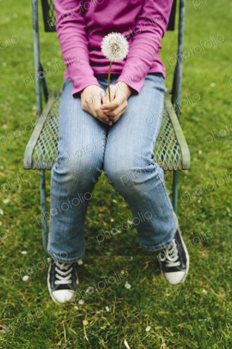A ten year old girl sitting in chair on lush, green grass, holding a dandelion clock…