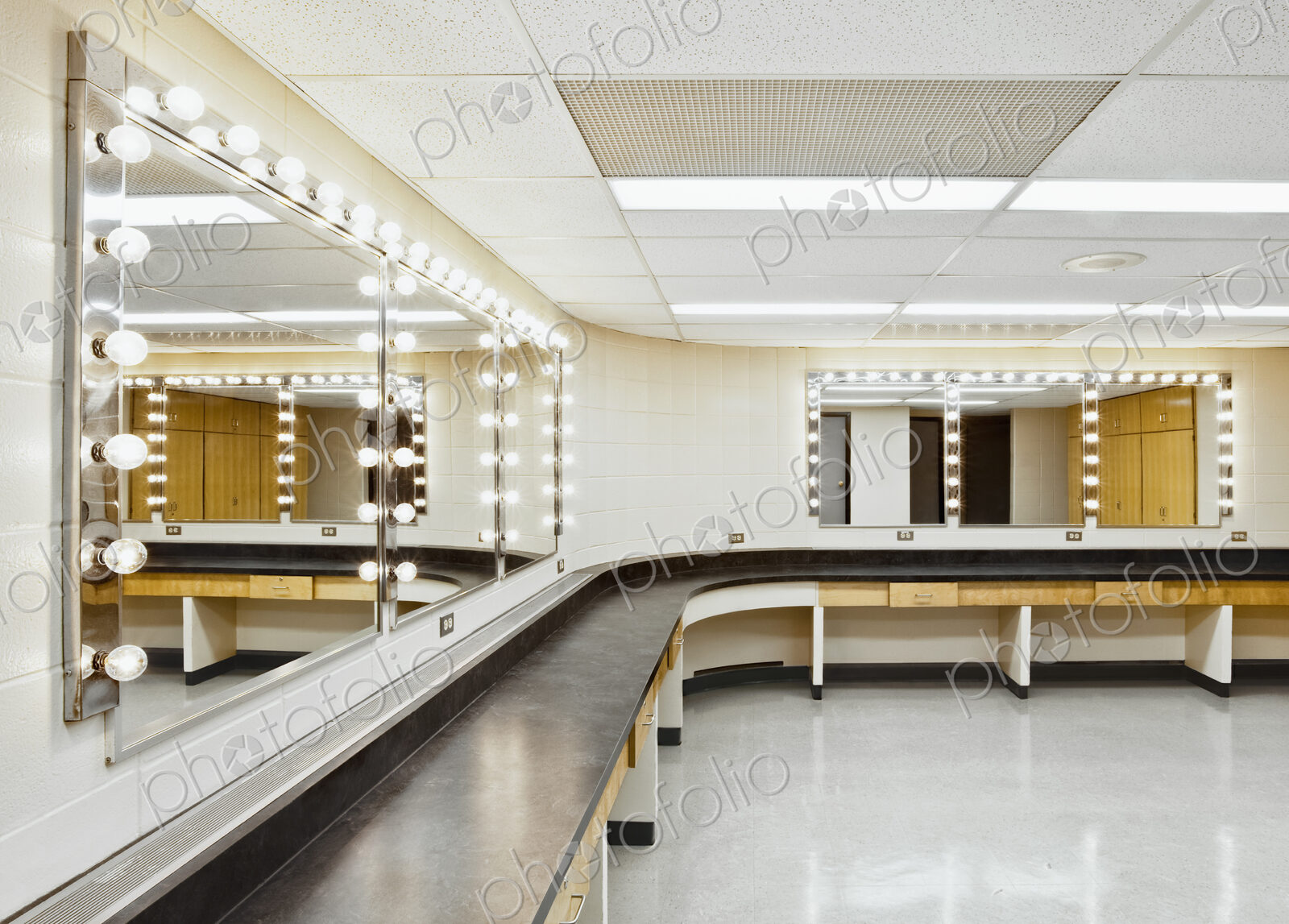 A theatre dressing room with a long worktop and seats and big mirrors with bulb…