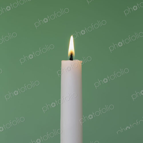 A thin white wax candle with a small lit flame with a green background.