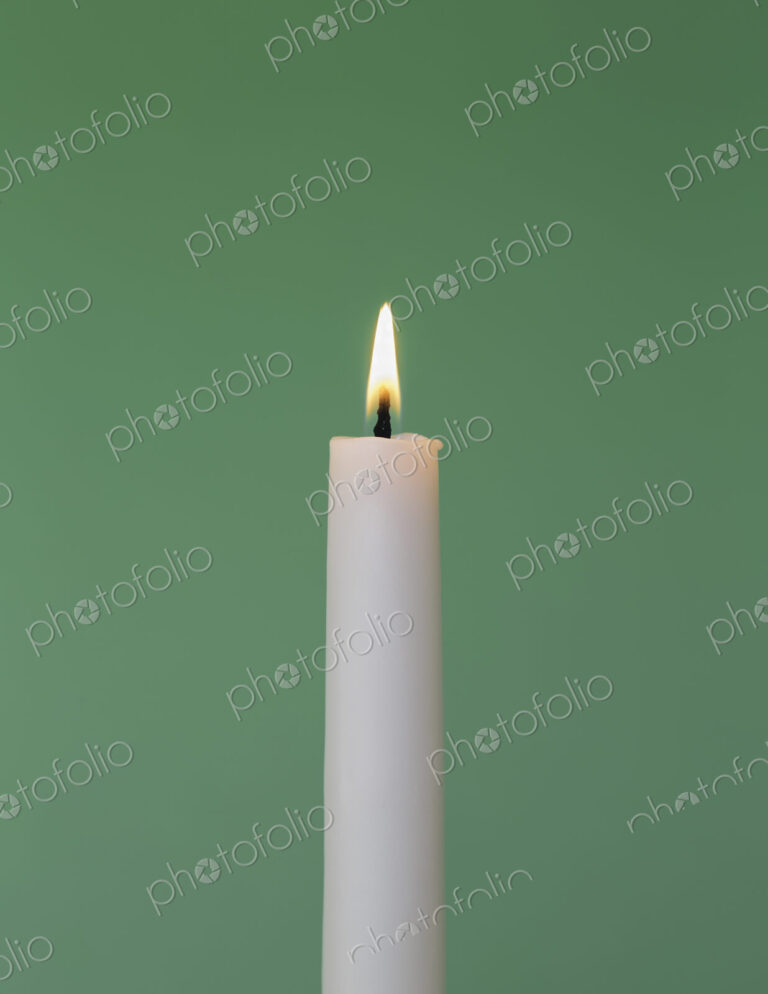 A thin white wax candle with a small lit flame with a green background.