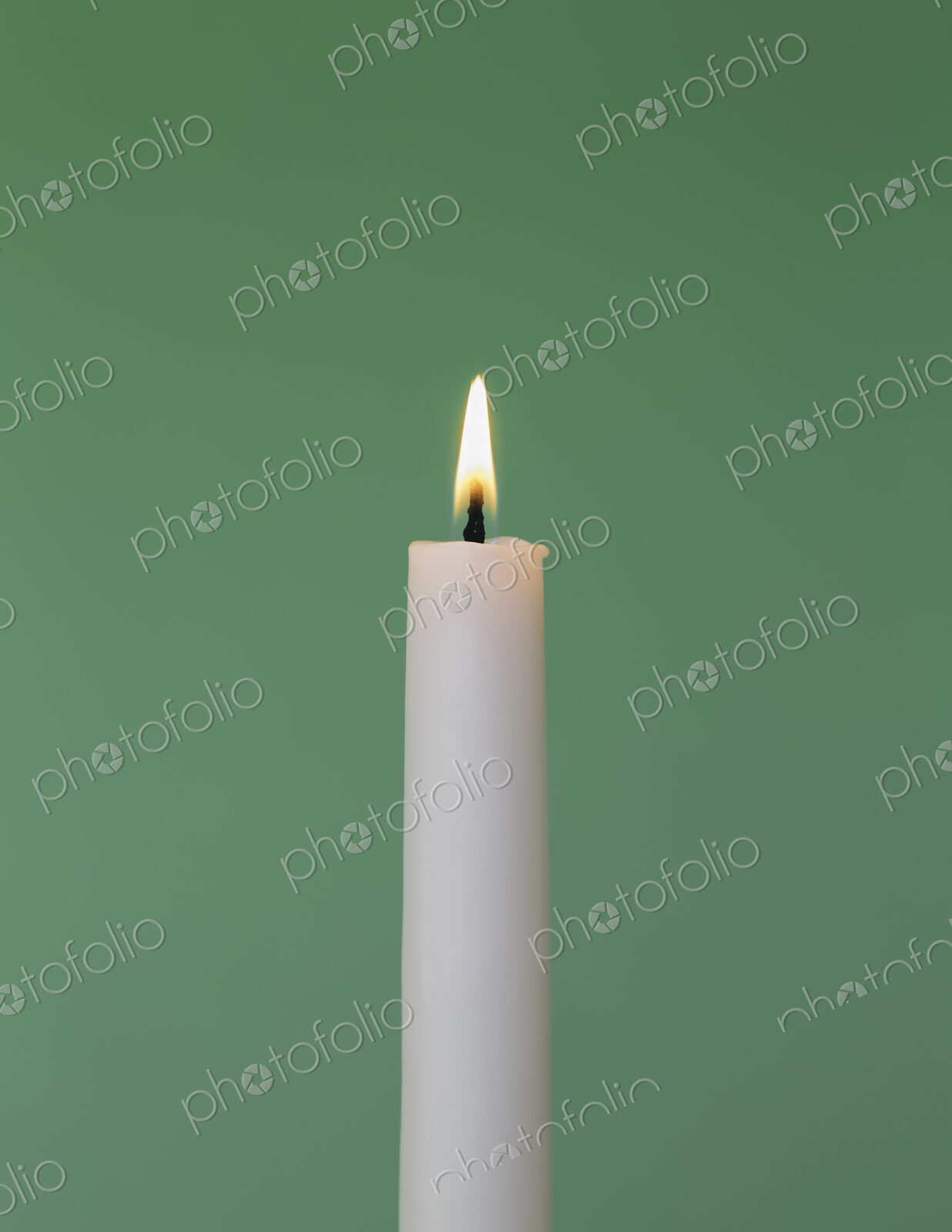 A thin white wax candle with a small lit flame with a green background.
