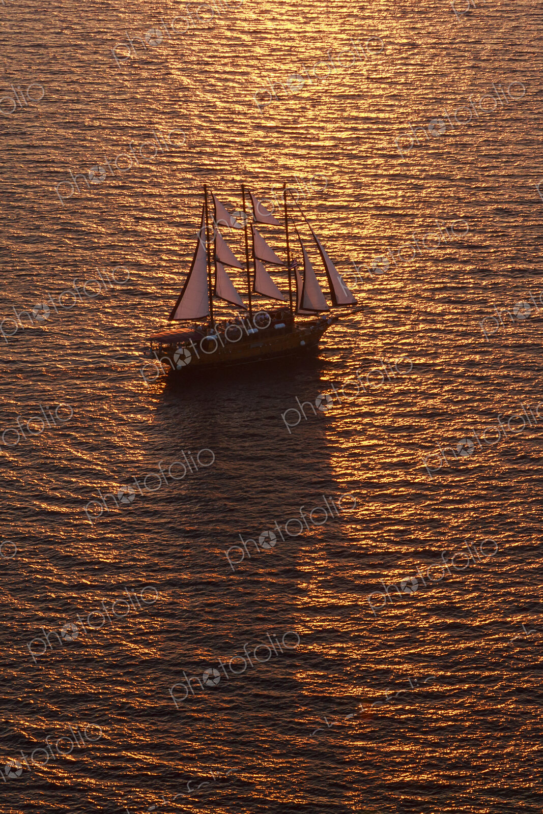 A three-masted sailing ship with full sail on the Aegean Sea at sunset.