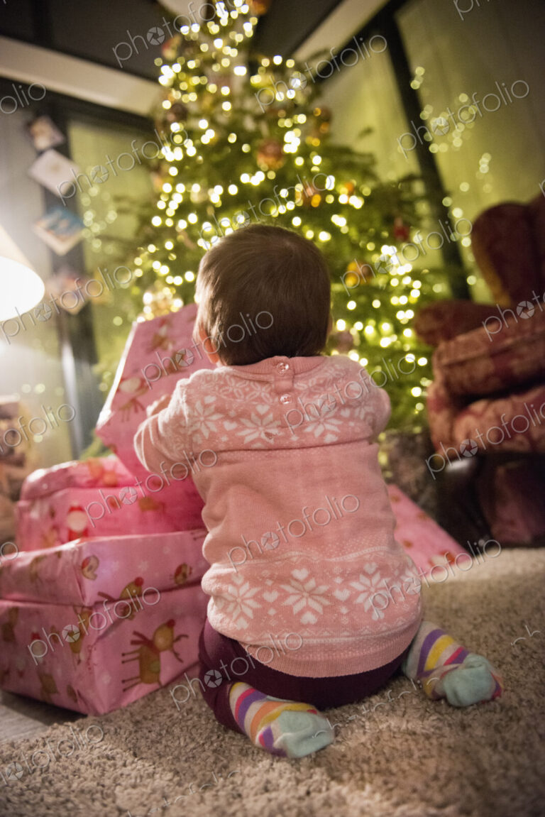 A toddler beside a pile of presents under a Christmas tree.