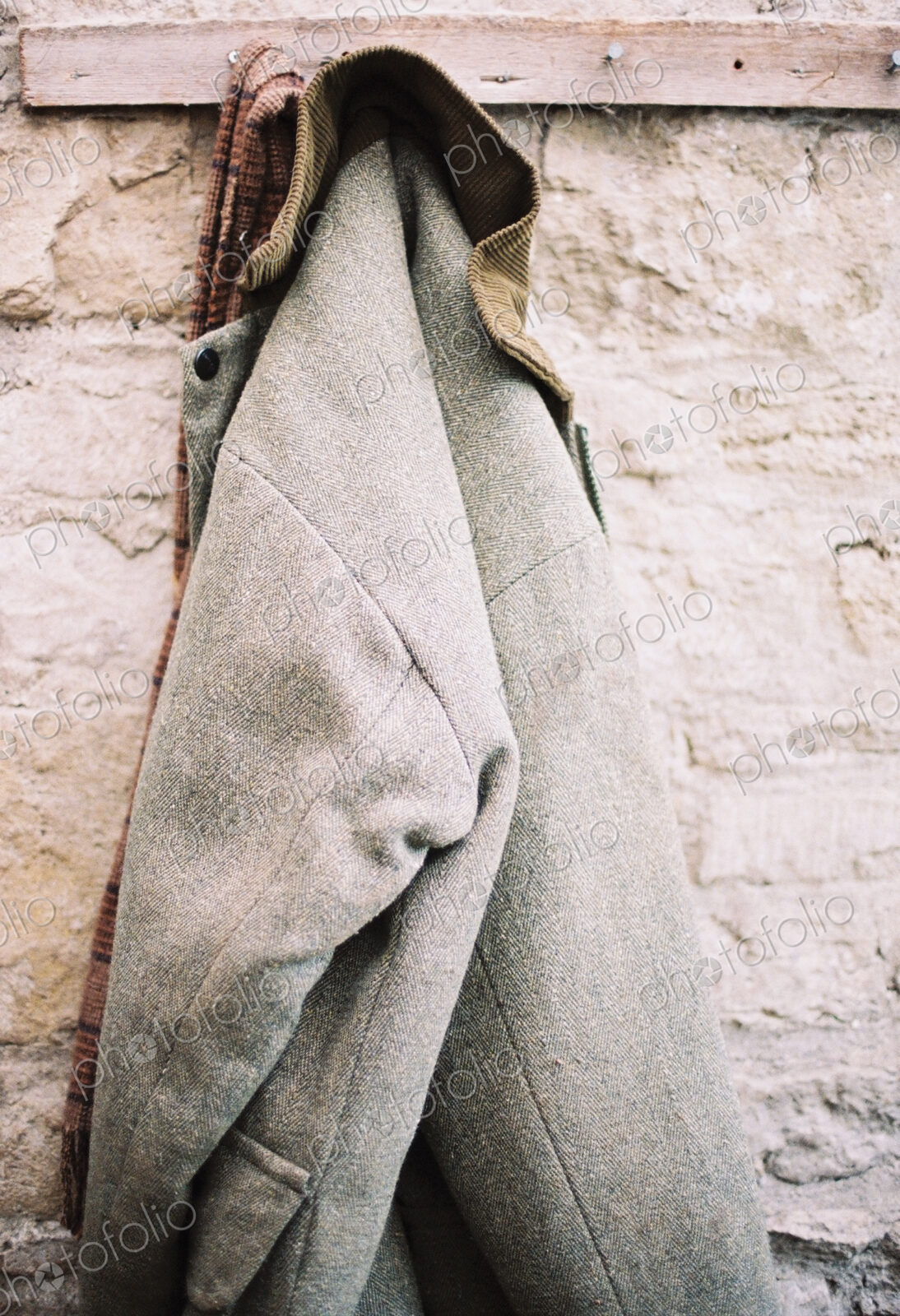 A traditional warm jacket with corduroy collar hanging from a pen on a stone wall.
