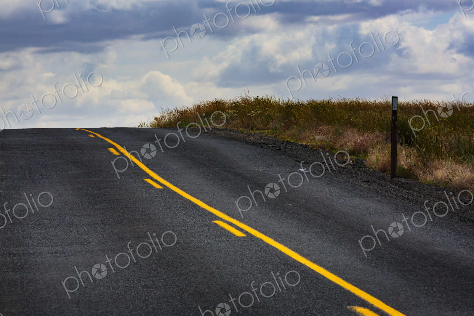 A two lane road with yellow central line, one solid and one broken through countryside.