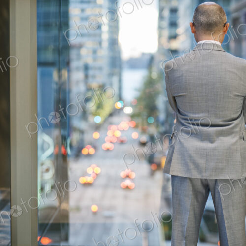 A view from behind a businessman standing at a window looking out at a city street… A view from behind a businessman standing at a window looking out at a city street…