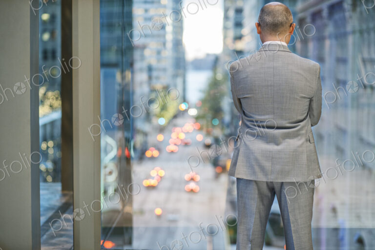 A view from behind a businessman standing at a window looking out at a city street…