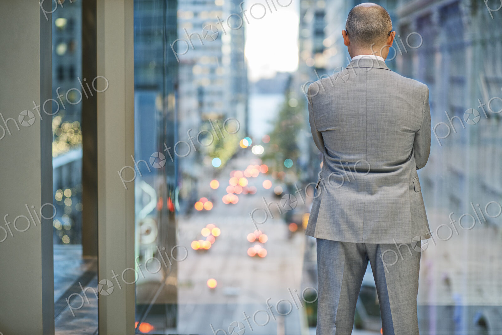 A view from behind a businessman standing at a window looking out at a city street…