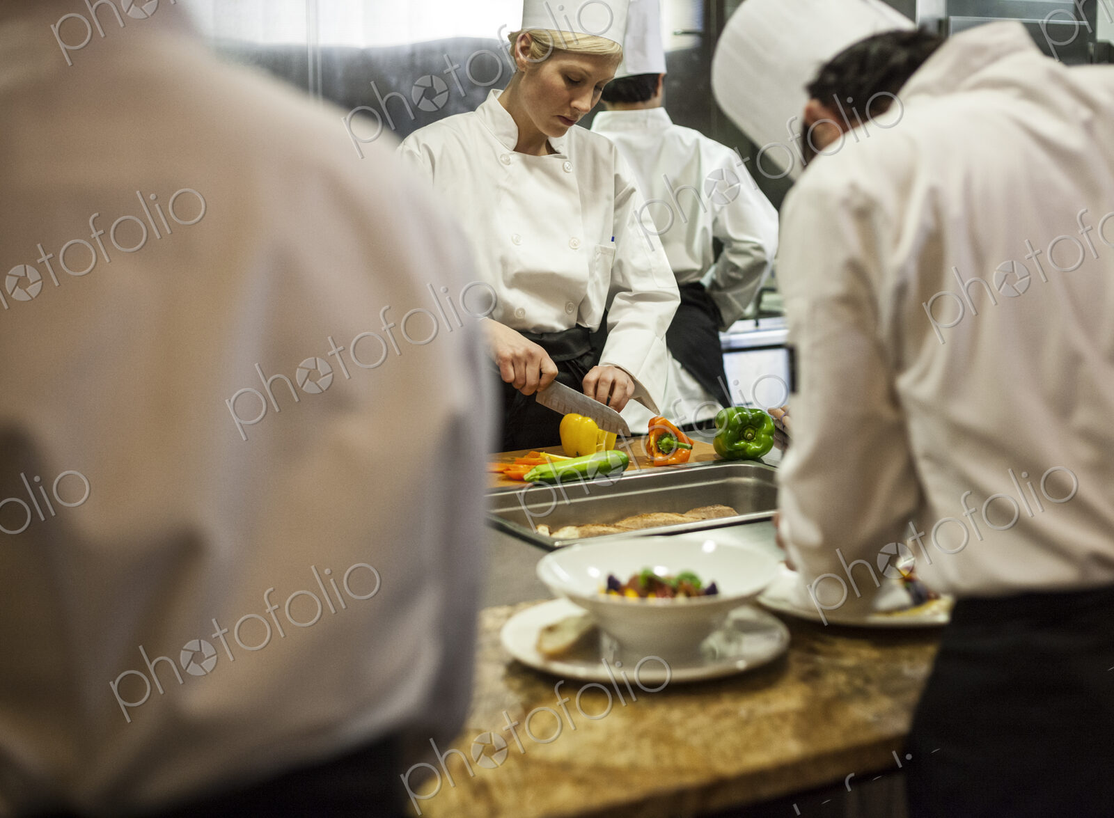 A view looking through kitchen employees of a female chef cutting vegetables.