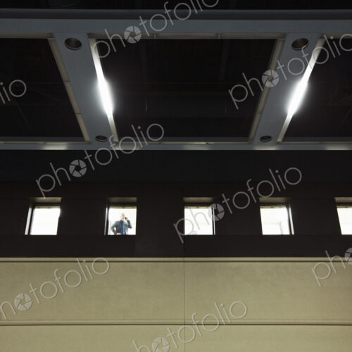 A view looking up at a businessman standing at an interior office window talking on a… A view looking up at a businessman standing at an interior office window talking on a…