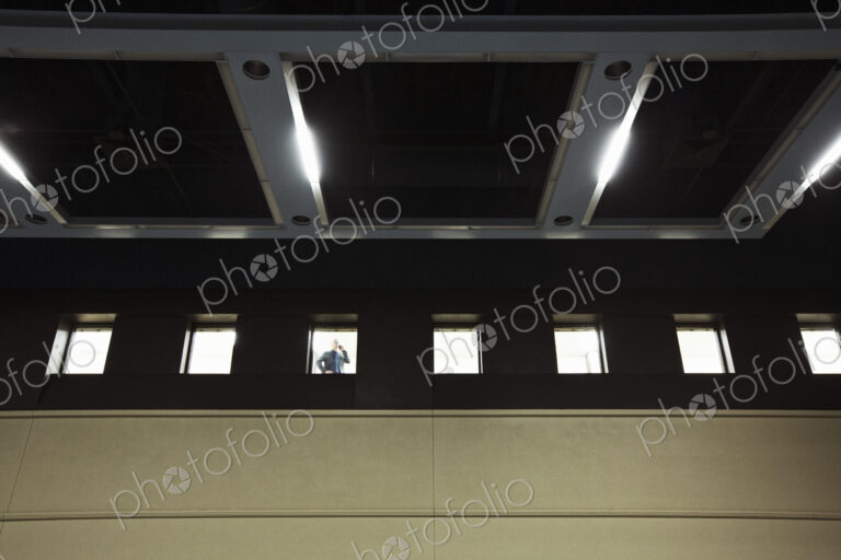 A view looking up at a businessman standing at an interior office window talking on a…