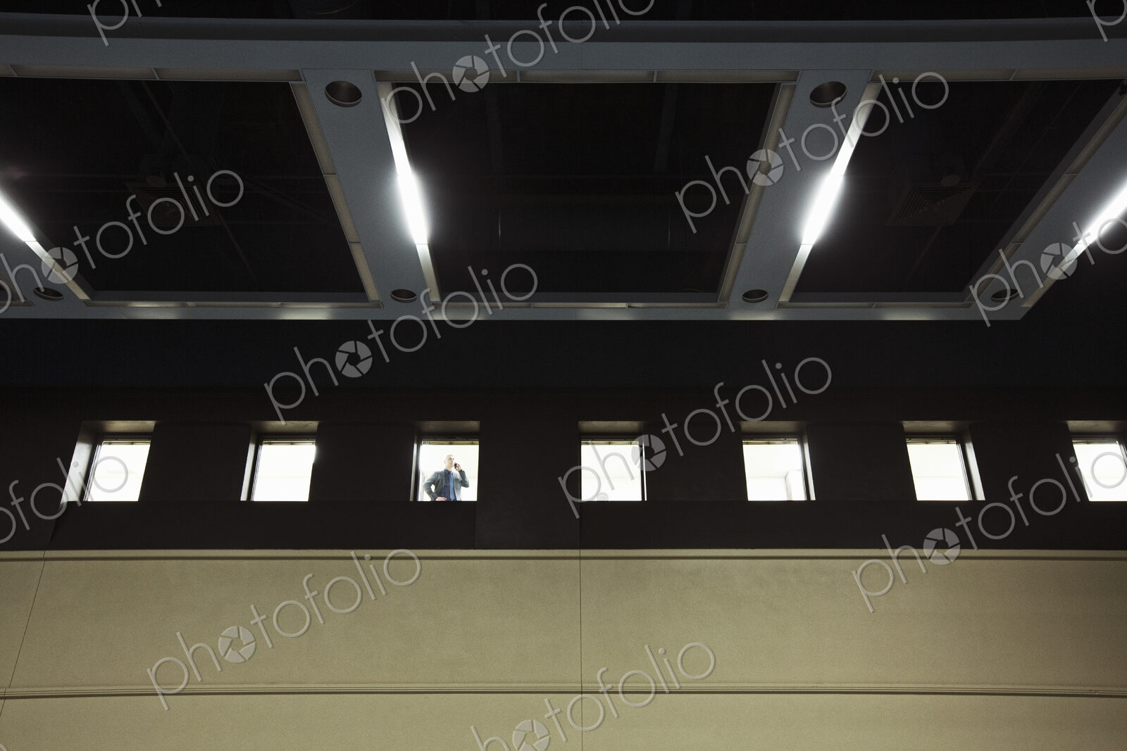 A view looking up at a businessman standing at an interior office window talking on a…
