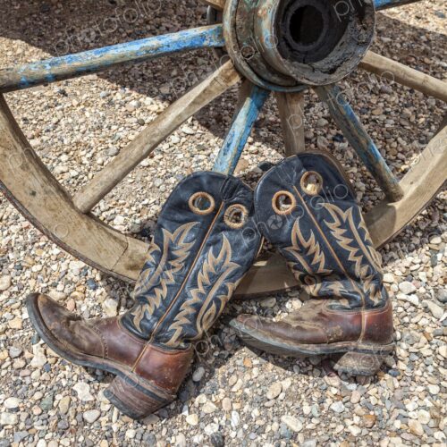 A weathered pair of cowboy boots rest against an old wooden wagon wheel on gravel,… A weathered pair of cowboy boots rest against an old wooden wagon wheel on gravel,…