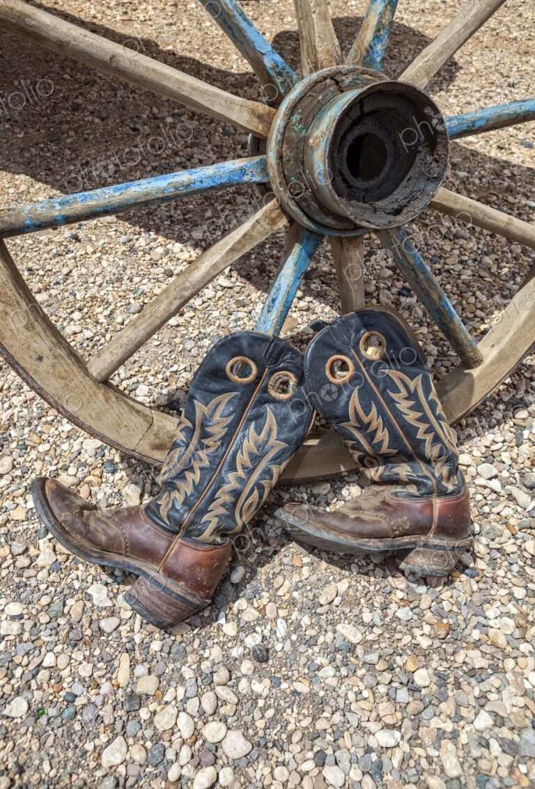 A weathered pair of cowboy boots rest against an old wooden wagon wheel on gravel,…
