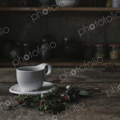 A white china cup and saucer on a kitchen table and a twig of pine and berries. A white china cup and saucer on a kitchen table and a twig of pine and berries.